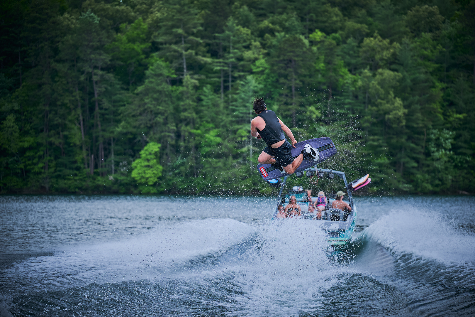 Wakeboarder spins mid-air above a powerful Axis wake with forested hills in the background.