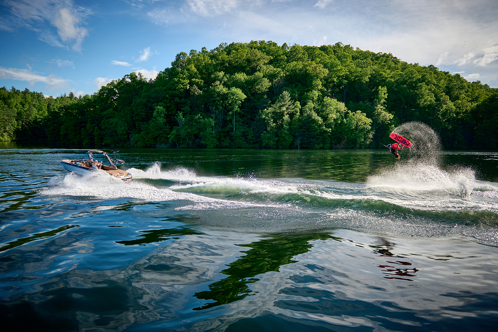 Wakeboarder gets airborne behind a Malibu 21 LX, framed by calm water and trees.